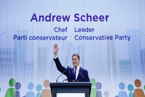 Andrew Scheer, der Vorsitzende der konservativen Partei, steht vor einem Podium mit zwei Mikrofonen und einem Bildschirm, mit einem Banner im Hintergrund mit Text und Gebäudeabbildungen.