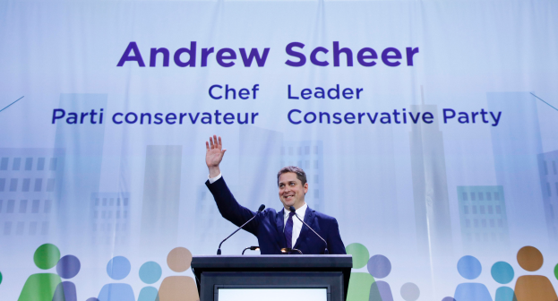Andrew Scheer, der Vorsitzende der konservativen Partei, steht vor einem Podium mit zwei Mikrofonen und einem Bildschirm, mit einem Banner im Hintergrund mit Text und Gebäudeabbildungen.