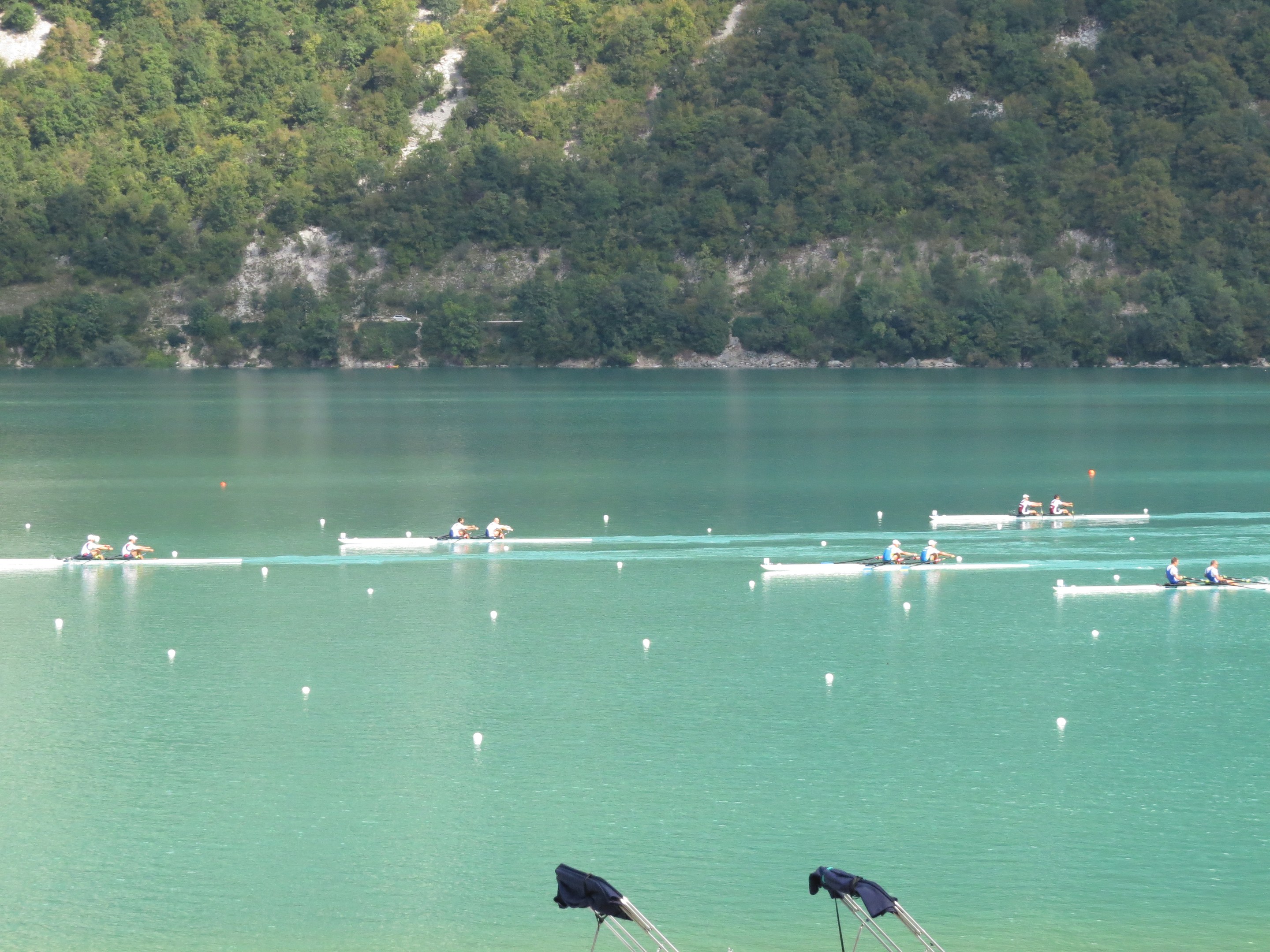 Eine Gruppe von Menschen, die in einem Wettbewerb auf einem Gewässer mit Bäumen und einem Hügel im Hintergrund rudern, und Kleiderstände unten sichtbar sind.