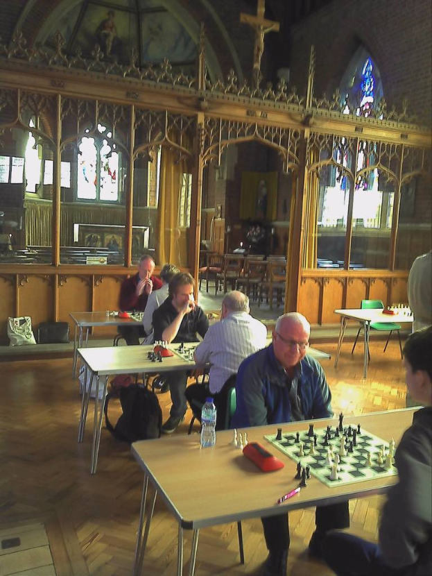 Eine Gruppe von Menschen, die Schach an Tischen in einer Kirche spielen, mit einem Mann in einem blauen Hemd, der eine Flasche und einen Stift auf seinem Tisch hat, und einem Glasfenster mit einem Kreuzsymbol im Hintergrund.