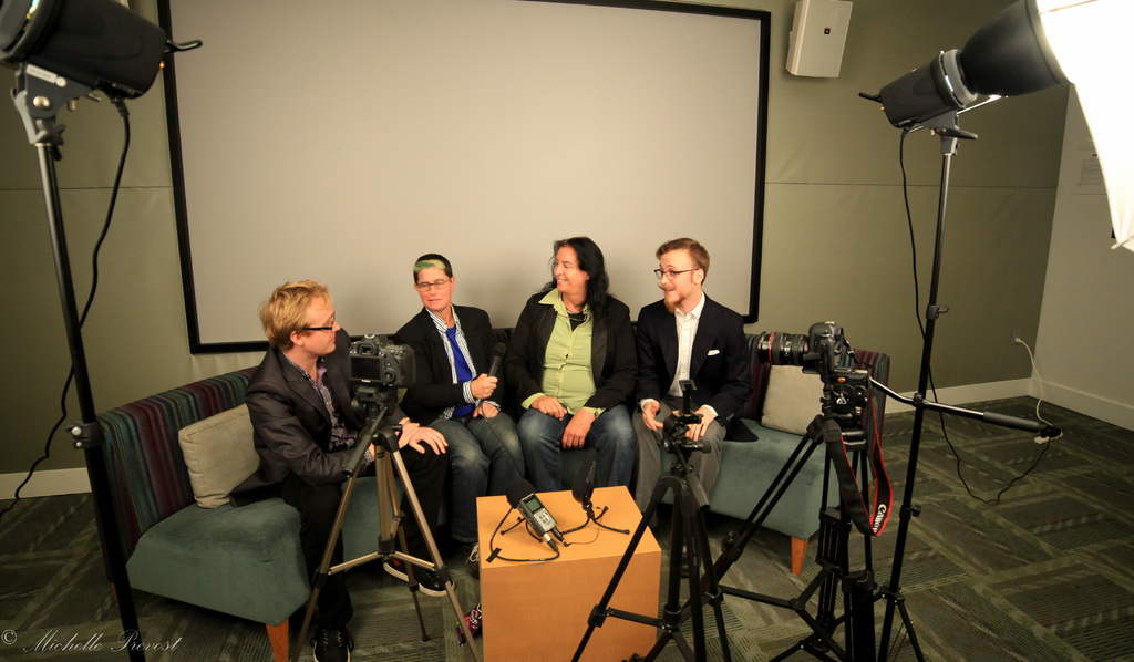 Eine Gruppe von drei Personen sitzt auf einem Sofa und gibt ein Interview, wobei eine Person ein Mikrofon hält, ein Aufnahmegerät auf dem Tisch in der Mitte steht, Kameras auf beiden Seiten und ein Projektionsschirm dahinter zu sehen ist.