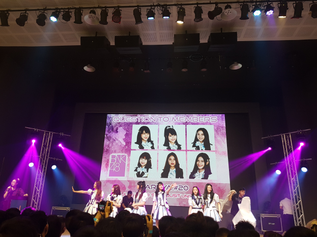 Eine Gruppe von Menschen, die auf einer hell erleuchteten Bühne performen, mit einer Menge darunter und einem Menschen mit Mikrofon und einem anderen, der ein Instrument spielt, sowie einem großen Bildschirm im Hintergrund, der Bilder von BK48 zeigt.