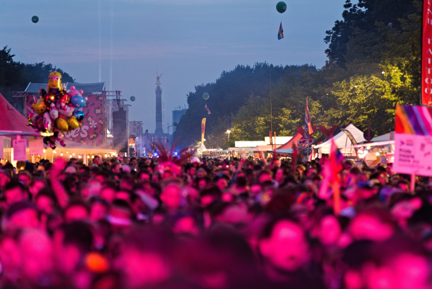 Große Menschenmenge auf einem nächtlichen Musikfestival mit bunten Lichtern, Ballons, Zelten, Bannern, Fahnen, Bäumen, einem Turm und einem sternenklaren Himmel im Hintergrund.