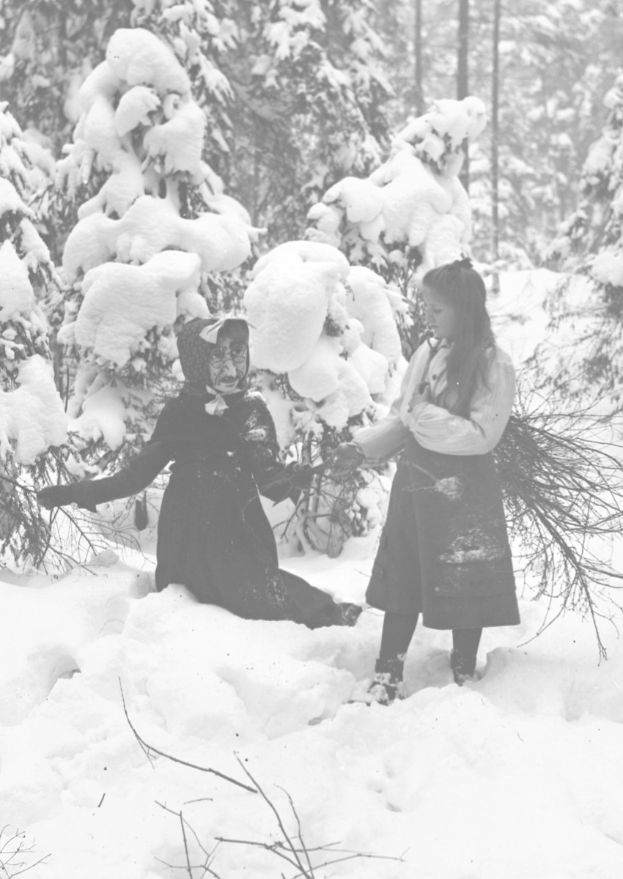 Zwei Kinder beim Spielen in einem schneebedeckten Waldgebiet, umgeben von schneebedeckten Bäumen, auf einem Schwarz-Weiß-Foto.
