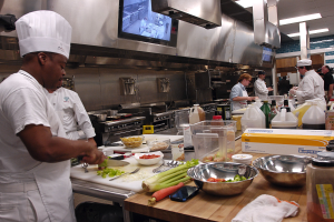 Eine Gruppe von Köchen in einer Küche, die Essen auf einem Schneidbrett zubereiten, wobei ein Koch im Vordergrund eine weiße Schürze und eine Mütze trägt und ein Messer hält und von verschiedenen Utensilien, Schüsseln, Gemüse, einem Mobiltelefon, Flaschen und anderen Gegenständen umgeben ist, mit einem Fernseher und Deckenlampen im Hintergrund.