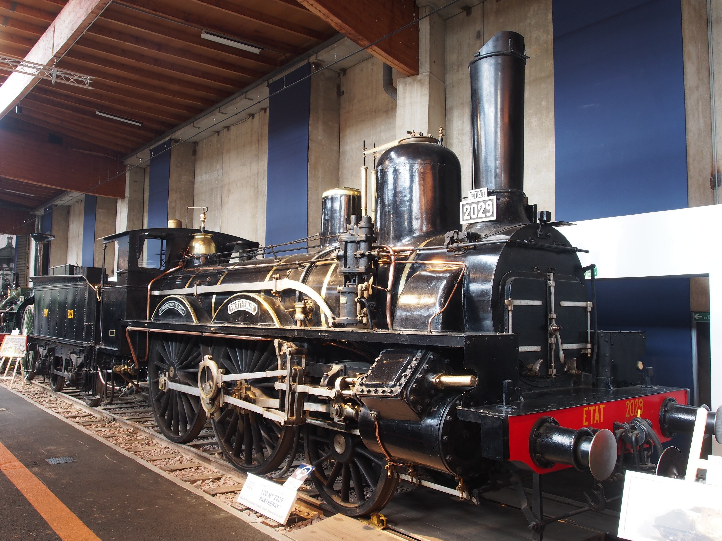 Alte Dampflokomotive in Museums-Atmosphäre mit umliegenden Schienensträngen, Informationsschildern und anderen Ausstellungsstücken, vor einer Wand mit Deckenbeleuchtung.
