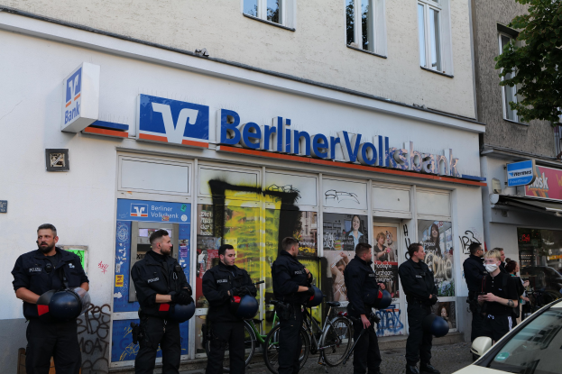 Eine Gruppe von Polizeibeamten steht vor einer Berliner Volksbank, einige halten Helme in den Händen, rechts ein Auto und links ein Baum; im Hintergrund sind Gebäude mit Fenstern und Texttafeln zu sehen.