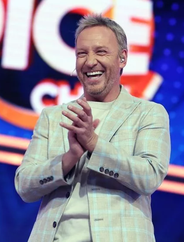 Ein Mann in einem Anzug steht auf einer Bühne, lächelt und klatscht, mit einem Schild im Hintergrund, auf dem 'The Voice' steht.