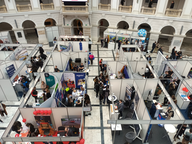 Große Gruppe von Menschen auf einer internationalen Fachmesse mit Ständen, Bannern, Tischen, Stühlen und Luftballons in einem Raum mit architektonischen Bögen, Säulen und einem Bildschirm im Hintergrund.