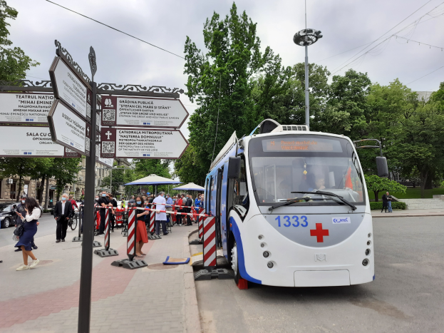 Ein weißer und blauer Bus parkt am Straßenrand in Serbien, umgeben von Menschen, mit Schildern, die einen Notfallbusdienst anzeigen, Absperrpoller, Fahrzeuge, ein Zelt, Pflanzen, Bäume, Drähte und ein Gebäude mit Fenstern unter einem bewölkten Himmel.