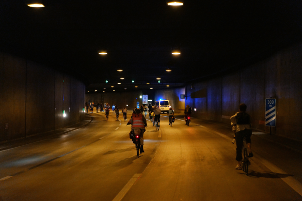 Eine Gruppe von Menschen, die nachts auf Fahrrädern eine Straße entlangfahren, beleuchtet von Lampen an der Decke, mit Wänden auf beiden Seiten und einem Fahrzeug im Hintergrund.