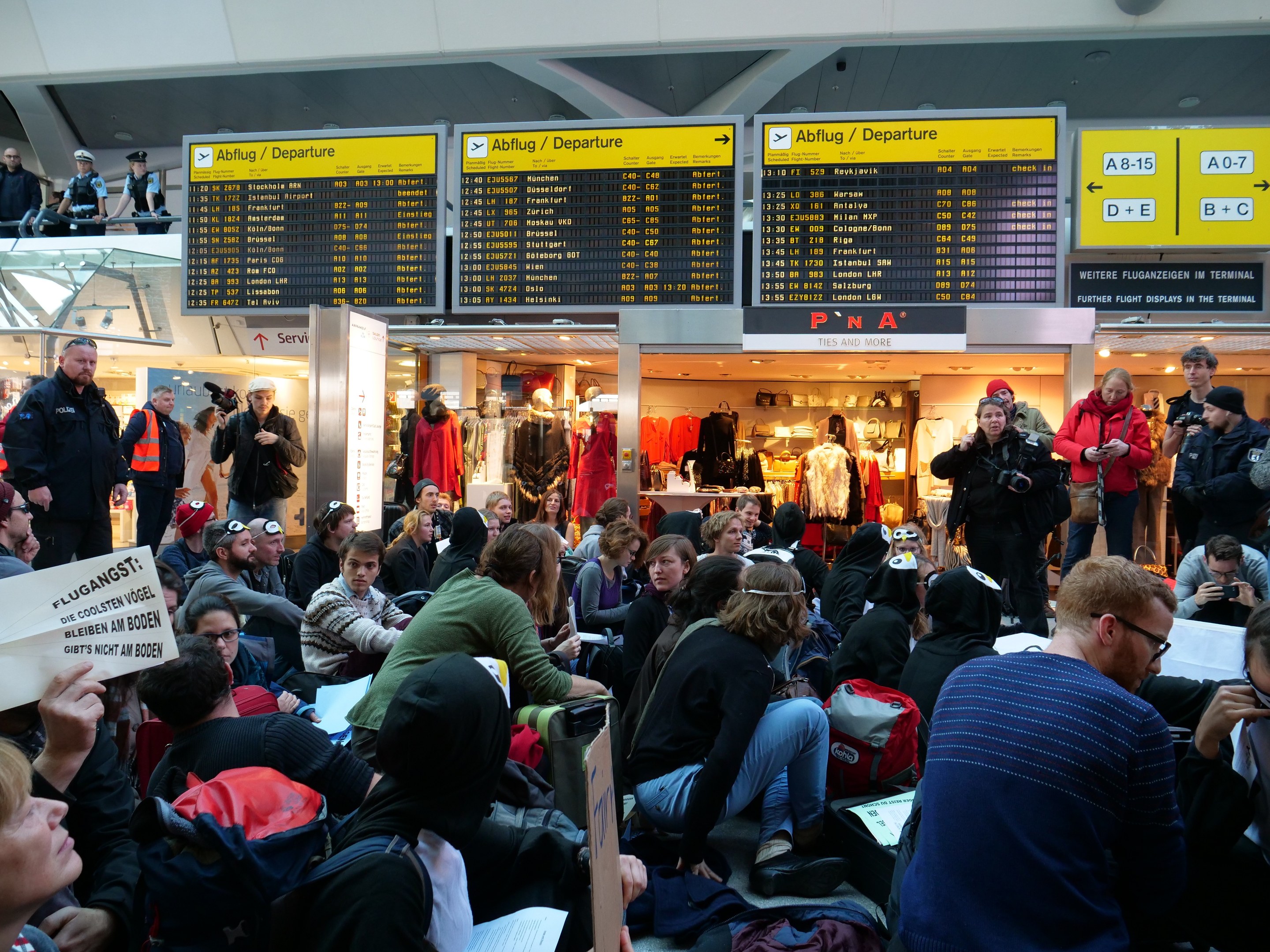 Eine große Gruppe von Menschen sitzt und steht in einem Flughafen während einer Demonstration, mit Schildern, Puppen und Deckenleuchten im Hintergrund.