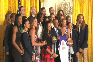 Präsident Obama und First Lady Michelle Obama posieren mit dem Damen-Basketball-Team im Oval Office des Weißen Hauses, halten einen Basketball, Pokal und lächeln in der Nähe einer Flagge, Vorhänge und einer Kerzenleuchter.