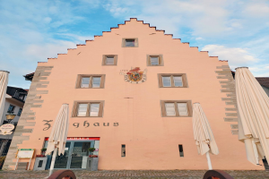 Ein rosafarbenes Gebäude mit einem zentralen Uhrenturm, umgeben von Fenstern, Schirmen, einem Schild, einer Straßenlaterne, Topfpflanzen unter einem bewölkten Himmel, repräsentiert München, Deutschland.