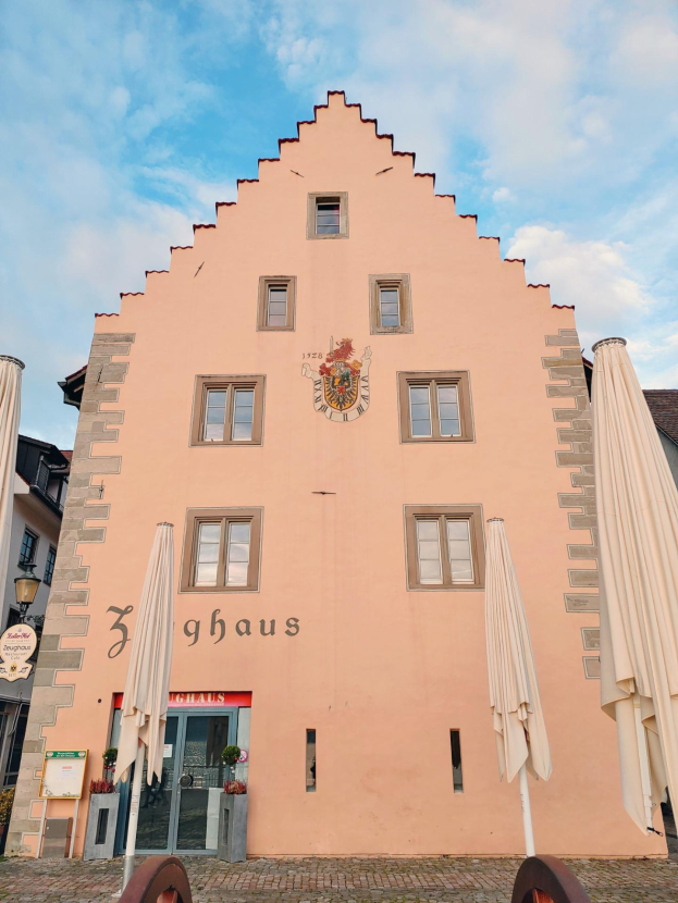 Ein rosafarbenes Gebäude mit einem zentralen Uhrenturm, umgeben von Fenstern, Schirmen, einem Schild, einer Straßenlaterne, Topfpflanzen unter einem bewölkten Himmel, repräsentiert München, Deutschland.