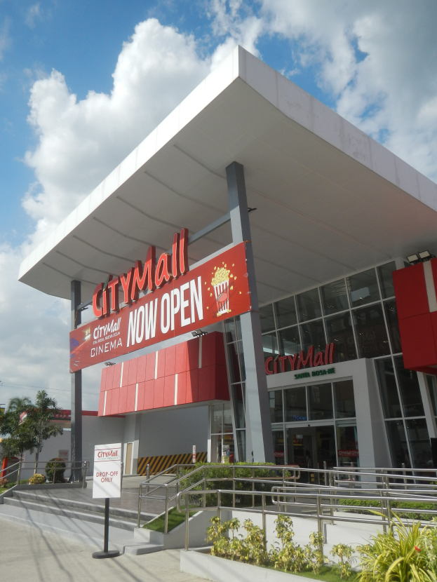 Ein belebter Stadtbummel mit einem Schild, auf dem "City Mall Now Open" steht, umgeben von Gebäuden, Schildern, Geländern, Grünflächen und Strommasten.