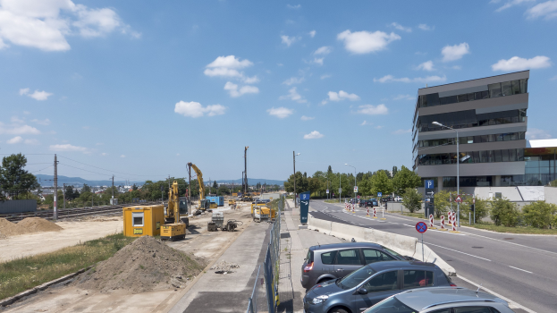 Eine Baustelle mit parkenden Autos, Gebäuden, Straßeninfrastruktur, Kränen, Bahnschienen und einem bewölkten Himmel im Hintergrund.