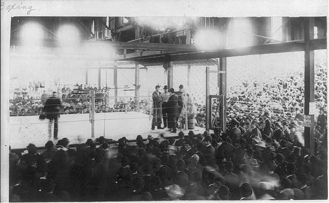 Schwarzes und weißes Foto einer Menge in einem Stadion, die einen Boxkampf beobachtet, mit einigen Menschen auf der Bühne und einigen im Publikum sitzend.