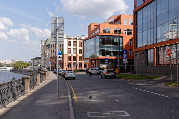 Stadtstraße mit parkenden Autos, Gebäuden, Schildern, Gras, einer Brücke über Wasser, Bäumen und einem bewölkten Himmel.