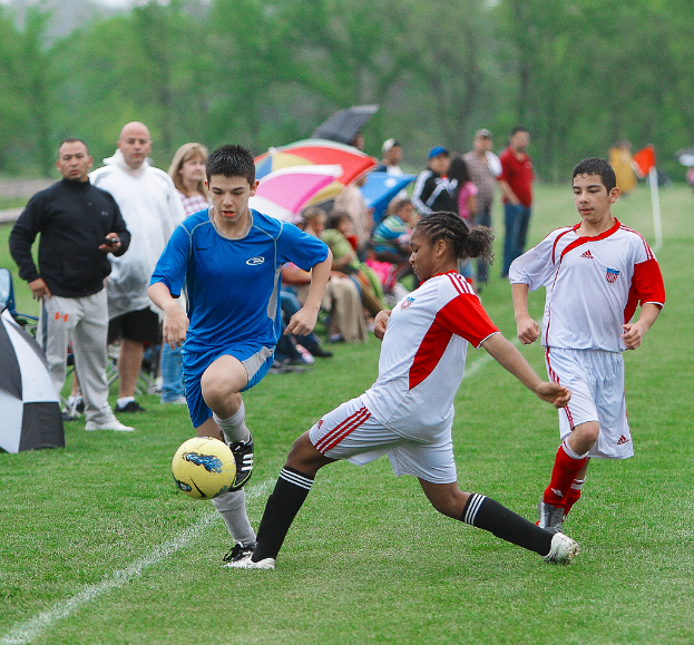 Drei Personen spielen Fußball mit einem gelben Ball, während eine Gruppe von Menschen mit Schirmen von hinten zusieht, mit Bäumen im Hintergrund.