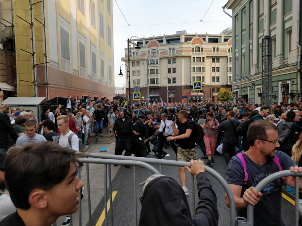 Eine große Menschenmenge steht hinter Metallabsperrungen auf einer Straße, einige halten Kameras, mit Gebäuden, Bäumen und Laternen im Hintergrund unter einem klaren Himmel.