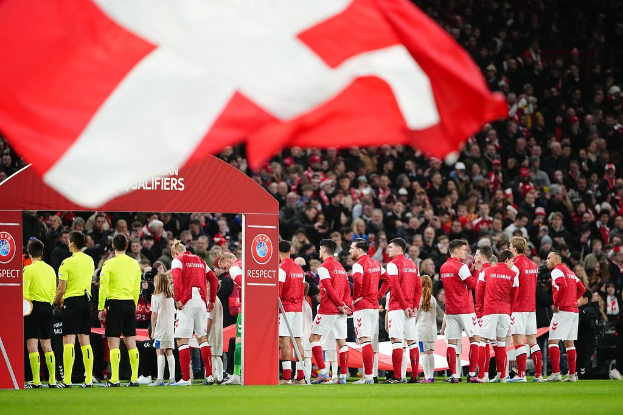 Eine Gruppe von Menschen auf einem Fußballfeld mit einer roten und weißen Flagge im Vordergrund, einem Bogen mit dem Text "Bayern München vs. Bayern München Wetten & Vorschau" im Hintergrund und einer großen Menge im Stadion.
