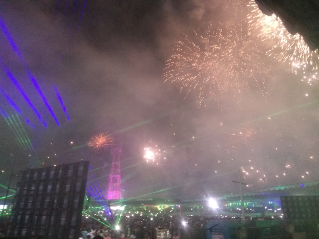 Eine Menschenmenge bei einem Konzert mit beleuchteten Gebäuden und Pfählen im Hintergrund, unter einem Himmel voller farbenfroher Feuerwerke.