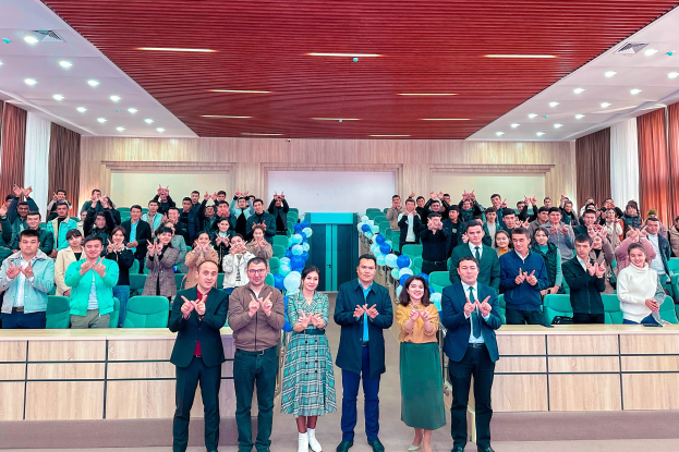 Eine Gruppe von Menschen steht auf einer Bühne vor einem Publikum, einige klatschen, mit Stühlen, Tischen, Ballons, Vorhängen und einer beleuchteten Decke im Hintergrund, was eine Schuleröffnung feierlich erscheinen lässt.