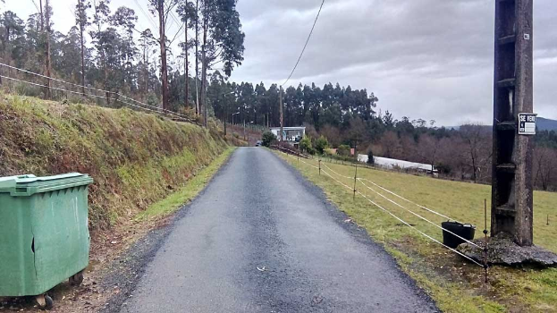 Ein grünter Müllbehälter sitzt neben einer von Gras, Pflanzen, Bäumen, Masten, Drähten und einem Zaun gesäumten Straße unter einem bewölktem Himmel, mit einem Wegweiser auf der rechten Seite und verstreuten Gegenständen auf der linken Seite, die den Tatort eines tödlichen Autounfalls markieren.