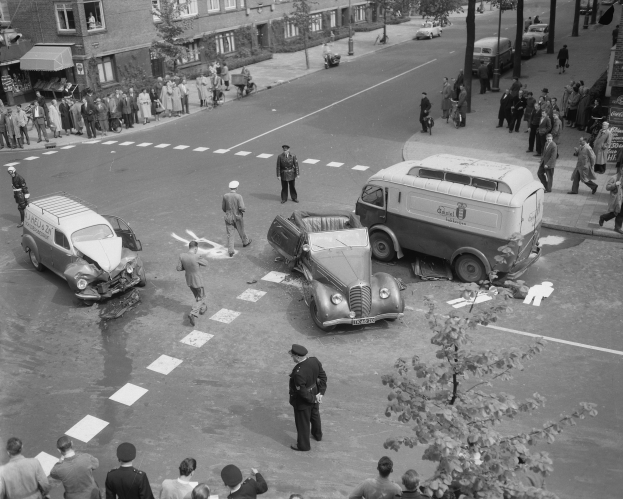 Schwarze und weiße Szenerie eines Autounfalls an einer Straßenkreuzung mit mehreren Fahrzeugen, darunter ein Van, und einer Gruppe von Schaulustigen; Gebäude, Bäume, Pfosten und Texttafeln sind im Hintergrund sichtbar.