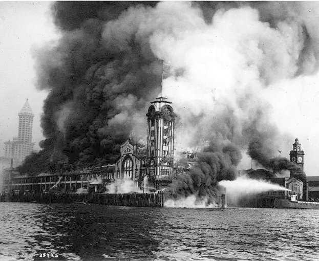 Schwarz-weißes Foto eines großen, von Flammen eingehüllten Gebäudes mit Rauch, der daraus aufsteigt, einem Gewässer im Vordergrund und anderen Gebäuden sowie dem Himmel im Hintergrund.