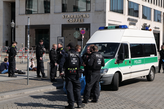 Eine Gruppe von Polizisten steht vor einem Starbucks-Kaffeehaus, mit einem Van auf der rechten Seite und einigen Menschen auf der linken Seite, vor einem Hintergrund mit Fenstern, einem Schild, einem Laternenpfahl und einem Zaun.
