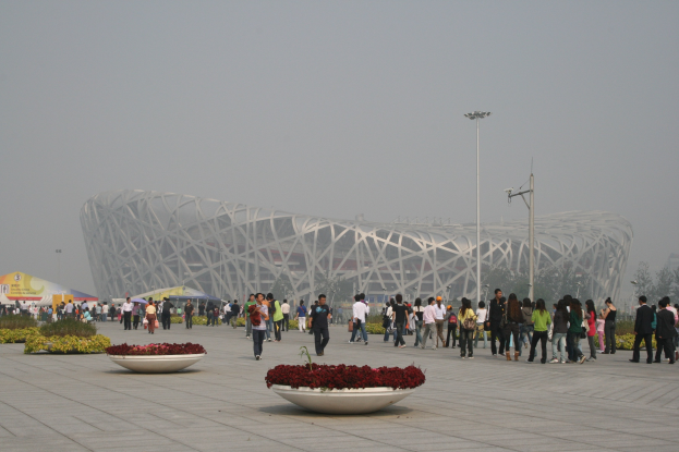 Olympisches Stadion in Peking mit Menschen, die herumlaufen, Pflanzen mit Blumen im Vordergrund, Laternenpfähle, Bäume und einen klaren blauen Himmel im Hintergrund.