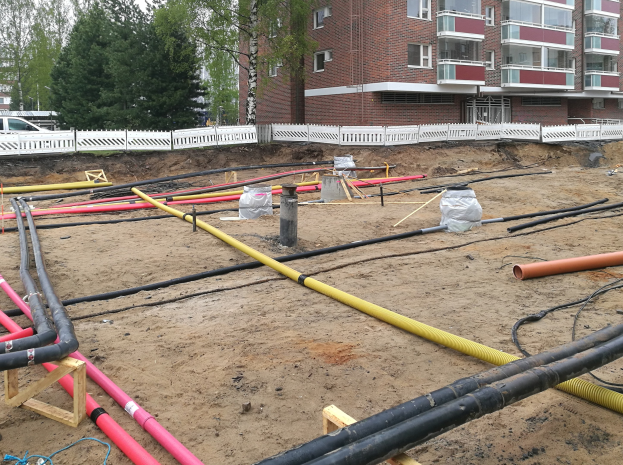 Großer Rohrstapel auf einer Baustelle mit Sand, Holzobjekten, Folienabdeckungen und anderen Materialien vor einem Gebäude, mit Bäumen, anderen Gebäuden, Fahrzeugen und einem klaren blauen Himmel im Hintergrund.