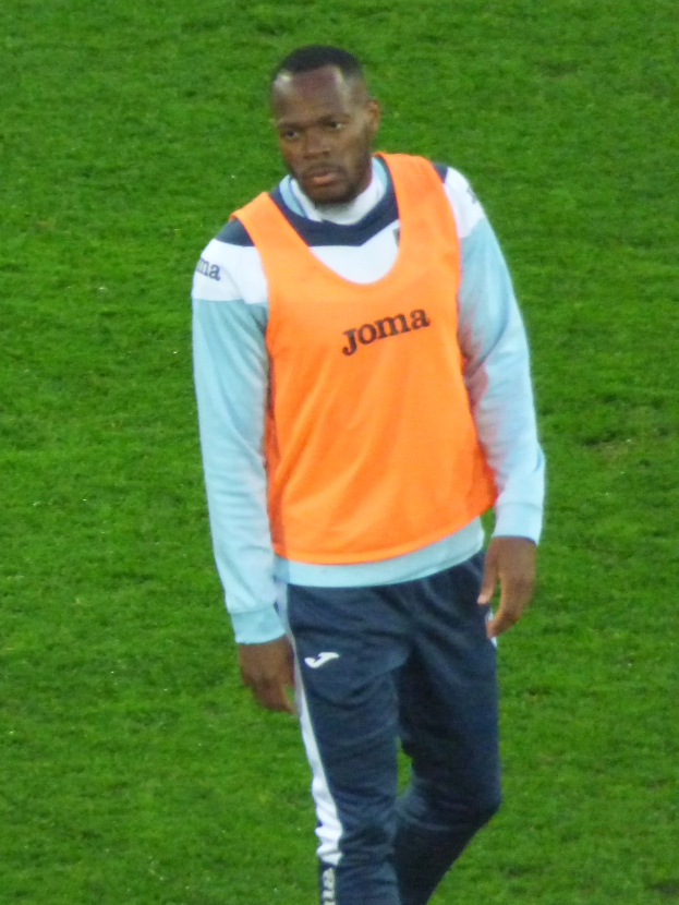 Ein Mann in einem orangen T-Shirt und blauen Hosen, der wie ein Fussballspieler aussieht, geht entschlossen über ein grünes Feld.