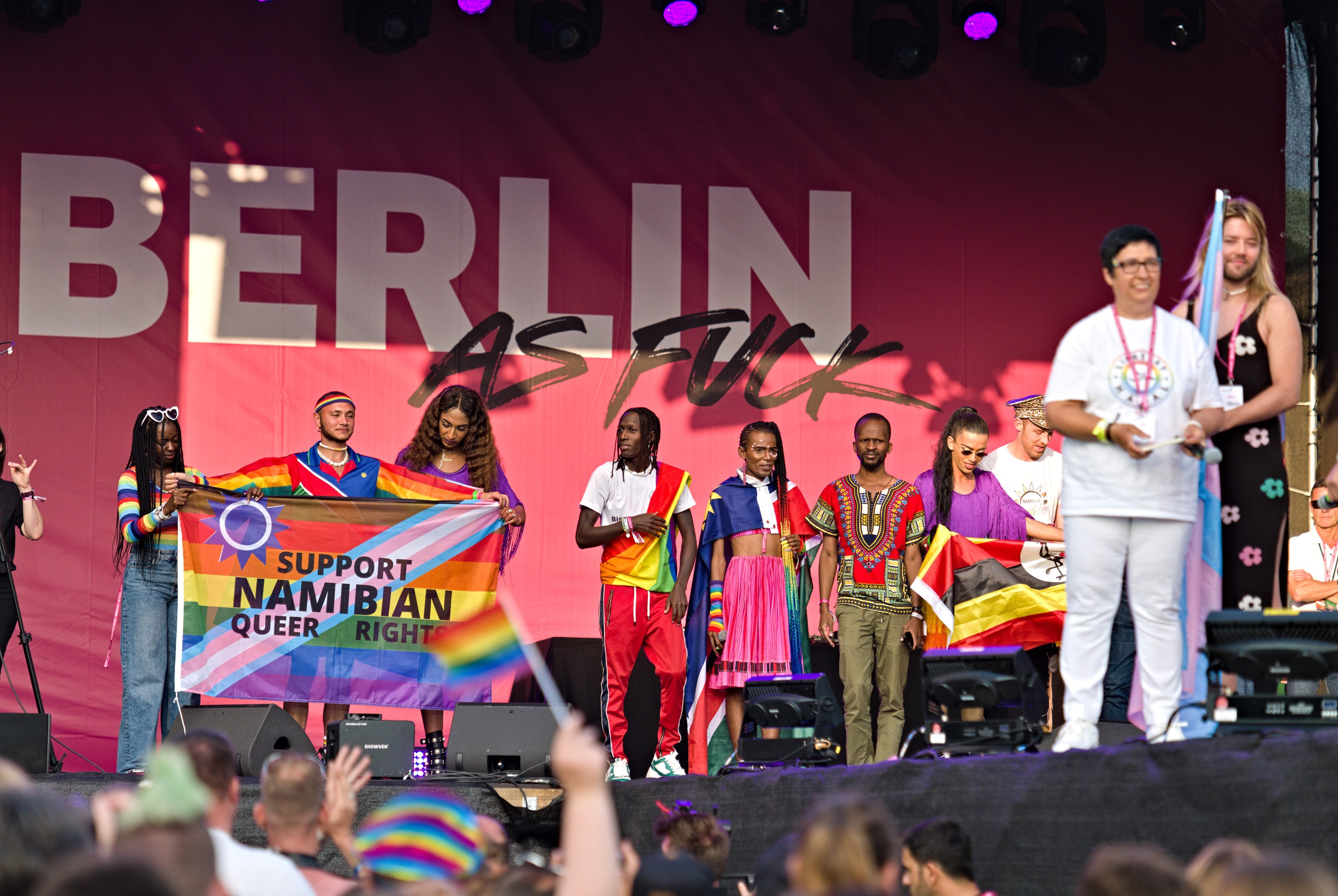 Leute auf einer Bühne mit einem Banner, auf dem 'Berlin as Fuck' steht, mit Lautsprechern, Mikrofonen und einer Menge unten, die ihre Hände hebt.