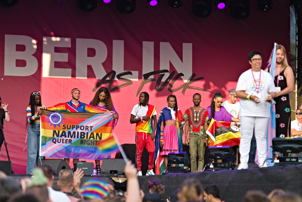Leute auf einer Bühne mit einem Banner, auf dem 'Berlin as Fuck' steht, mit Lautsprechern, Mikrofonen und einer Menge unten, die ihre Hände hebt.