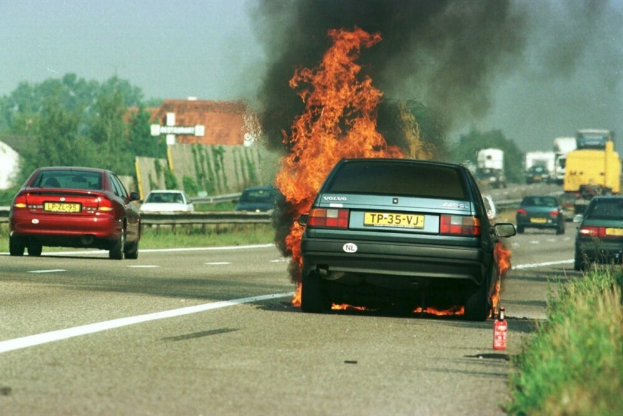 Ein Auto brennt am Straßenrand mit anderen Fahrzeugen in der Nähe, Tannen und Gebäude im Hintergrund unter einem klaren blauen Himmel und Gras mit einem Feuerlöscher auf der rechten Seite.