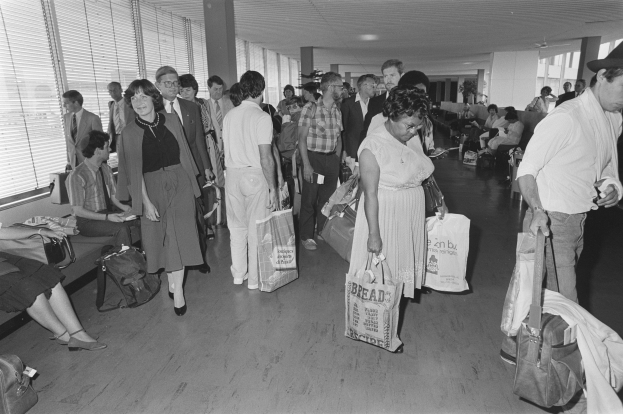 Schwarzes und weißes Bild von Reisenden in einem Flughafen, einige sitzen auf Bänken und andere stehen mit Gepäck, mit einem Mann und einer Frau, die Taschen halten, in der Mitte und Jalousien auf der linken Seite.