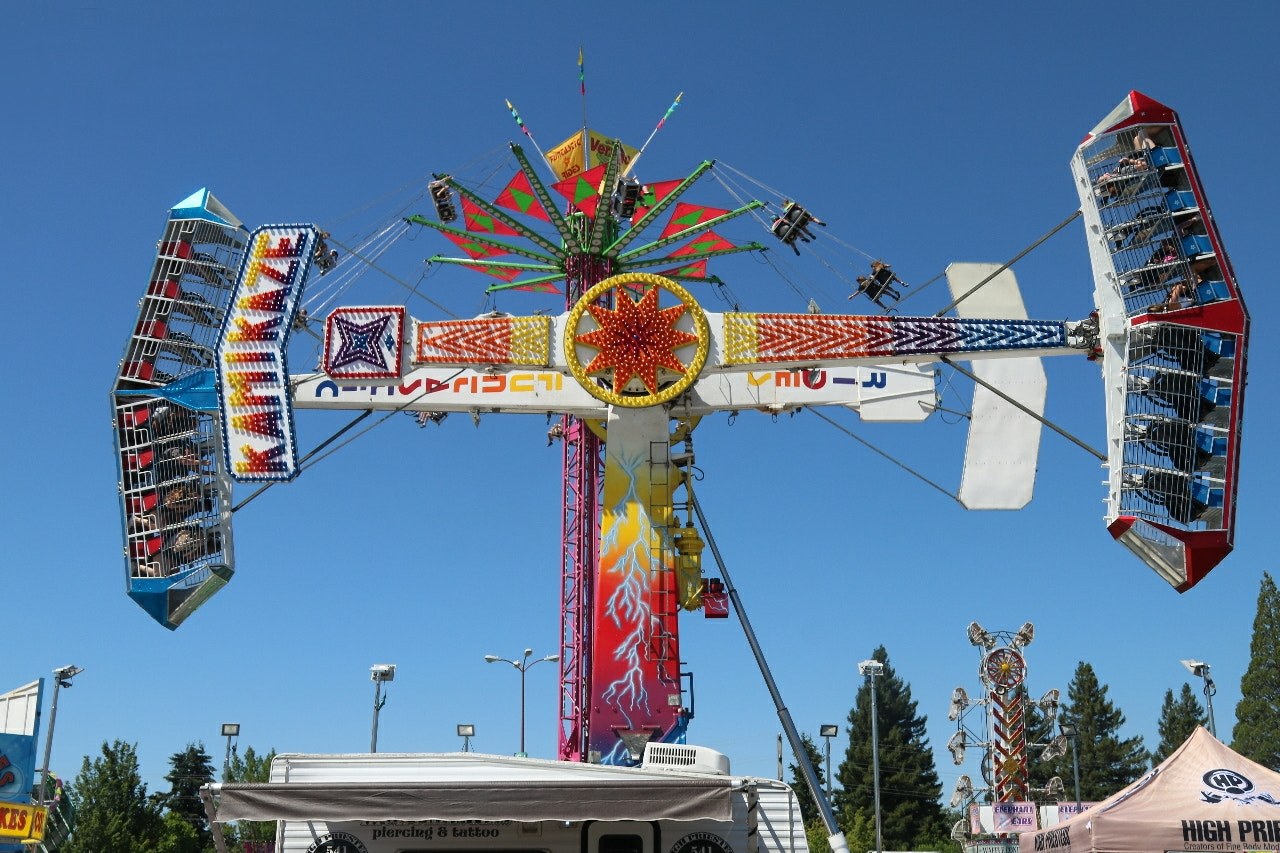 Ein buntes Karussell mit Passagieren, umgeben von Fahrbetrieben, Zelten, Bannern, Stangen und Lichtern, vor einem Himmel mit Bäumen.