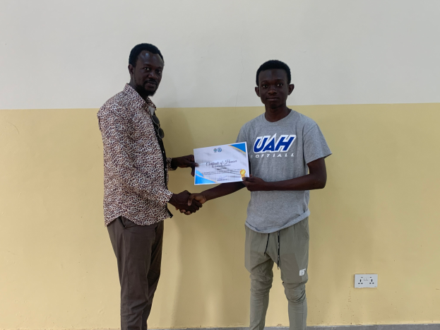 Zwei Männer schütteln sich vor einer gelben Wand die Hand, einer hält einen Stipendienschein der Universität Ghana.