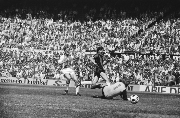 Eine Gruppe von Männern, die auf einem Feld Fußball spielen, mit Zuschauern und einem Banner mit der Aufschrift "Bundesliga 1978-79 Ajax 1-1 Hannover 96" im Hintergrund, in Schwarz-Weiß.