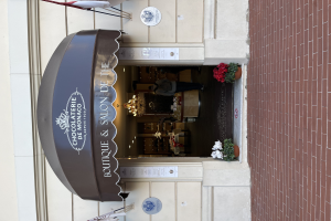 Ein Gebäude mit einer Tafel, auf der "Boutique & Salon de Monaco" steht, mit Außenbestuhlung, Blumentöpfen und einem Blick in das gut beleuchtete, luxuriöse Innere.