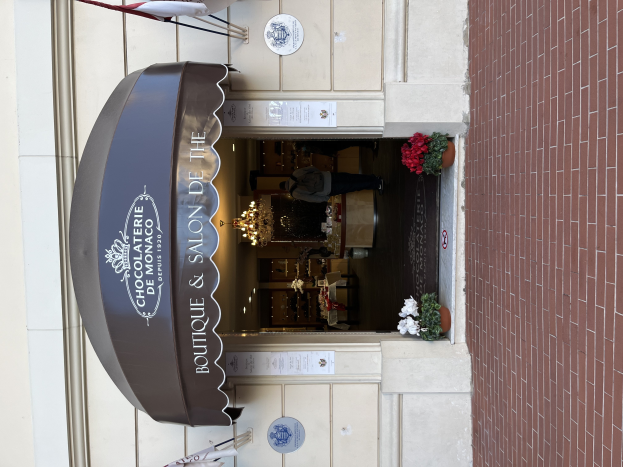 Ein Gebäude mit einer Tafel, auf der "Boutique & Salon de Monaco" steht, mit Außenbestuhlung, Blumentöpfen und einem Blick in das gut beleuchtete, luxuriöse Innere.