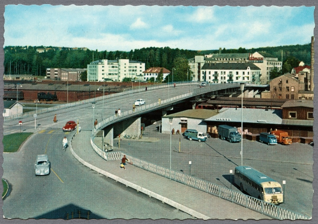 Altes Schwarz-Weiß-Foto einer Stadtstraße mit Fahrzeugen, Fußgängern auf einer Brücke, Gebäuden, Bäumen und einem bewölkten Himmel.