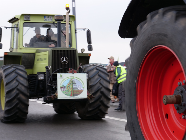 Grüner Traktor mit einem roten Rad, das eine Straße entlangfährt, umgeben von Menschen in Jacken und Mützen, mit Bäumen und einem klaren blauen Himmel im Hintergrund.