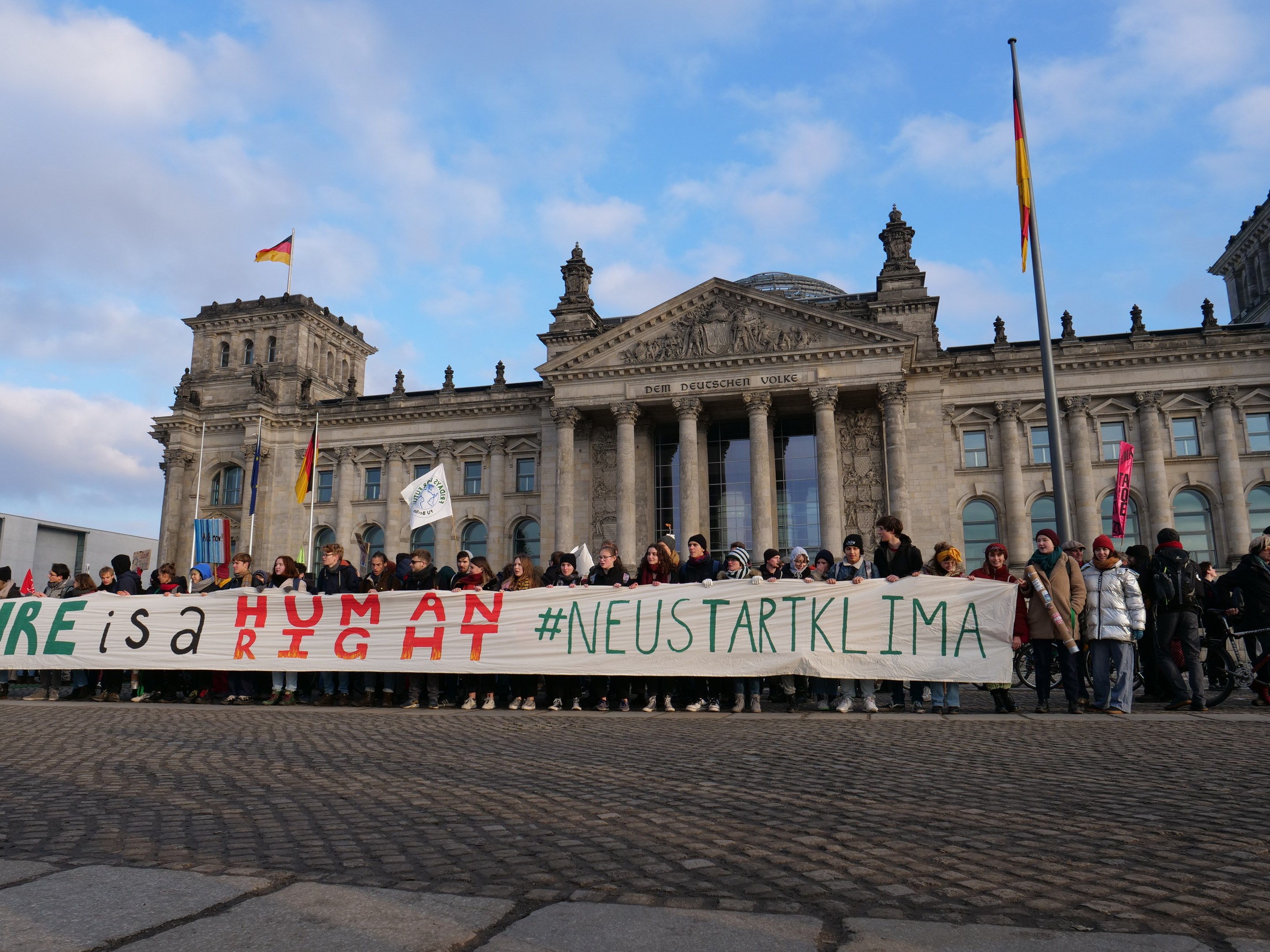 Eine Gruppe von Menschen steht vor dem Reichstaggebäude in Berlin und hält ein Banner mit der Aufschrift "Wir sind ein Menschenrecht" in den Händen.