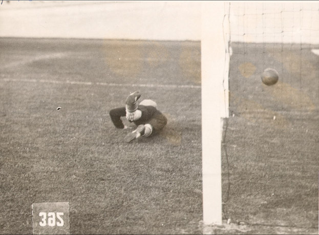 Ein Fussballspieler in Schwarz-Weiß, der einen Ball vor einem Torpfosten mit Netz auf der rechten Seite fängt, mit Text unten im Bild.