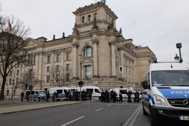 Eine Gruppe von Polizeibeamten steht vor dem Reichstag in Berlin, Deutschland, mit Fahrzeugen, einem Zaun, Verkehrszeichen, Laternen, Bäumen und Flaggen im Hintergrund, bei klarem Himmel.