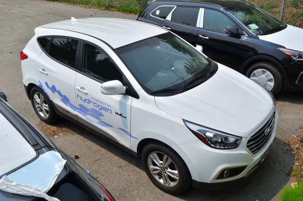 Drei Autos nebeneinander auf einem Parkplatz geparkt, wobei eines den Text Wasserstoff-getriebenes Fahrzeug anzeigt, ein Maschendrahtzaun im Hintergrund und Gras auf der rechten Seite.